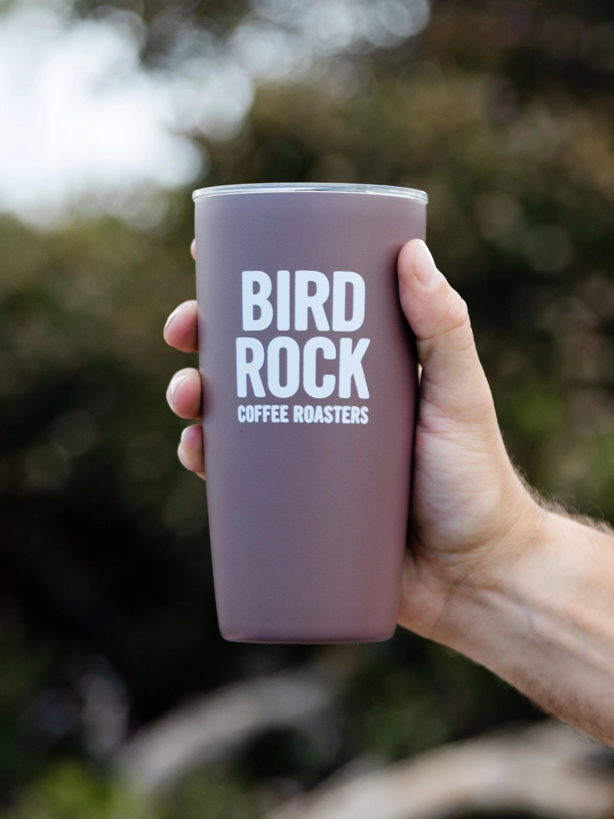A hand holding a matte mauve-taupe insulated tumbler with white text that reads 'BIRD ROCK COFFEE ROASTERS' against a blurred outdoor background.