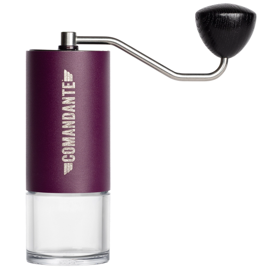 A manual coffee grinder with a metallic purple body, a clear glass lower chamber, a silver metal hand crank, and a black wooden handle knob. The brand name 'COMANDANTE' is printed vertically in white on the purple section.
