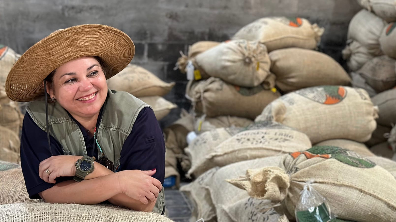 A smiling woman wearing a wide-brim straw hat and a green vest leans against a large burlap sack in a warehouse filled with stacked burlap bags.