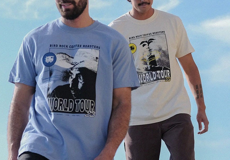 Two men wearing 'World Tour' t-shirts standing on a rocky coastal area with a scenic background.