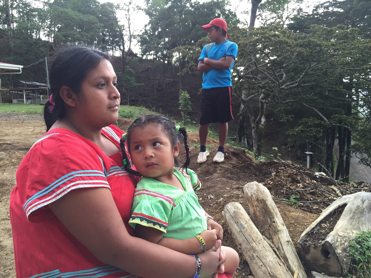 A woman in a traditional red dress holding a young girl in a green dress at Finca Auromar, a man in red cap behind