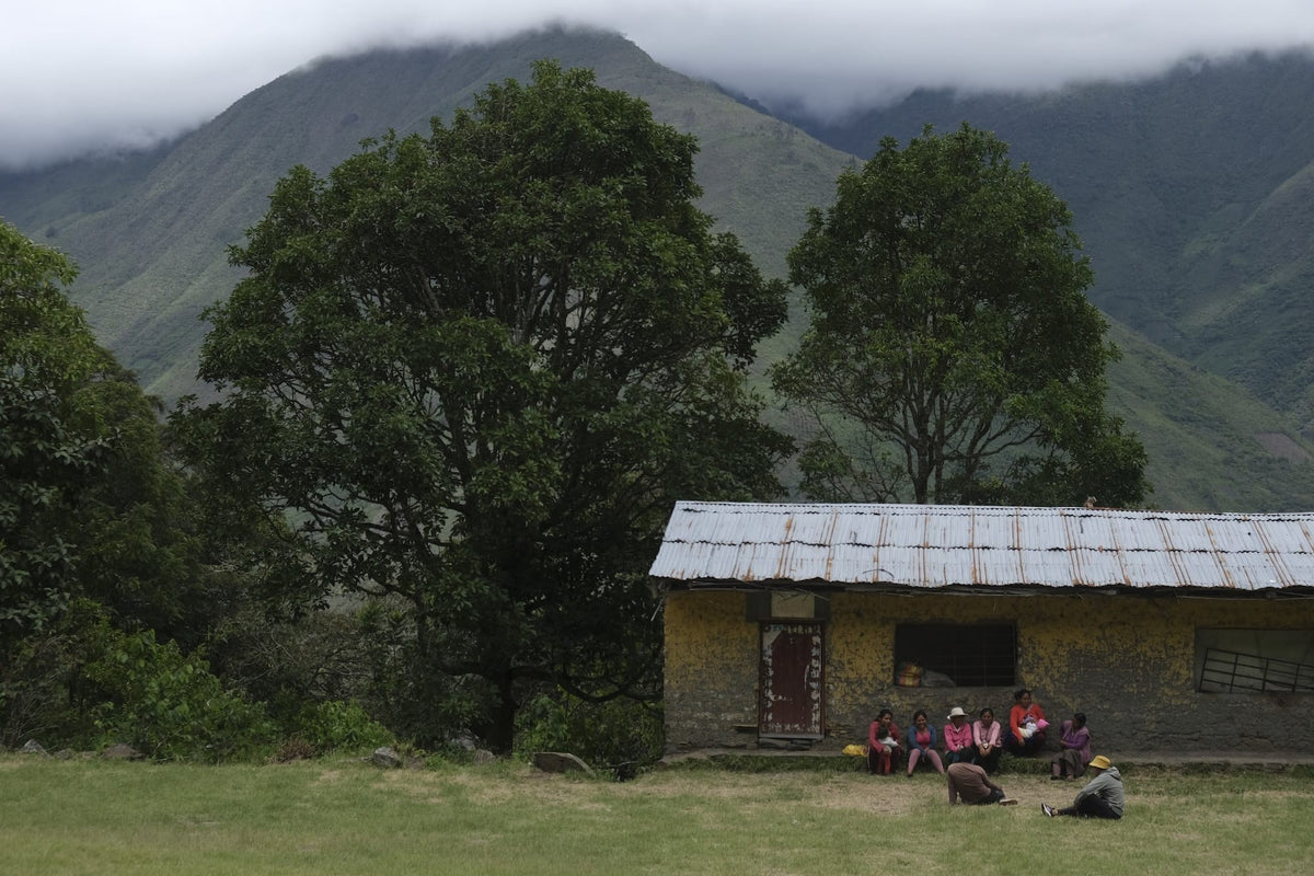 Cloud-capped green Andean mountains above a tin-roofed stone house, with a group of women seated in front